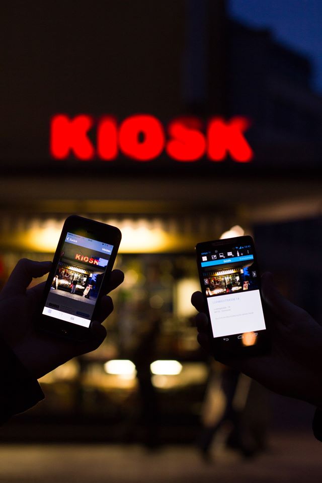 Ein nachts leuchtender Kiosk, vor dem zwei Menschen Handys mit der App halten